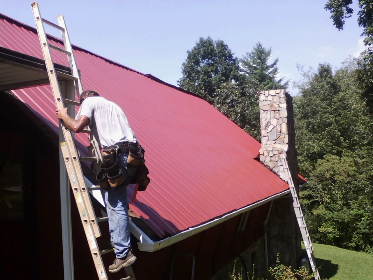 Roofer on a ladder beside a red metal roof and stone chimney — Soffit & Fascia Repair by Roofing Brothers in Cumberland Furnace