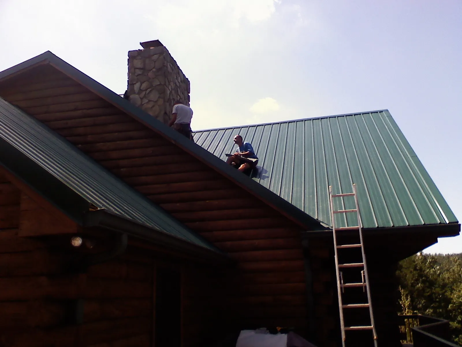 Roofers installing green metal panels beside a stone chimney on a log home in Cumberland Furnace